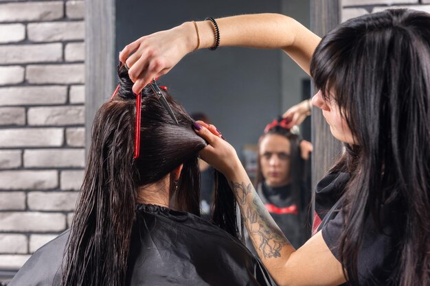 Close up female stylist with tattooed hands cutting and modeling brown hair by scissors and comb of young brunette woman while she is sitting in armchair in beauty salon