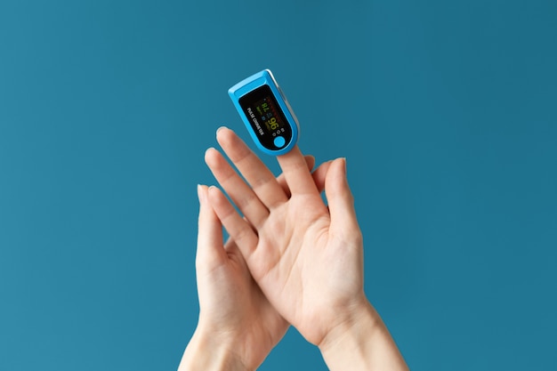 Close up of females hand with a pulse oximeter placed on the index finger. Blue background. The concept of measuring oxygen in the blood.