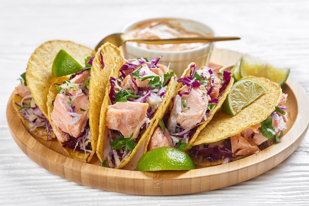 Close-up of fish tacos with red cabbage salad with spicy yogurt sauce sprinkled with finely chopped parsley served on a bamboo plate on a white wooden table, horizontal view from above