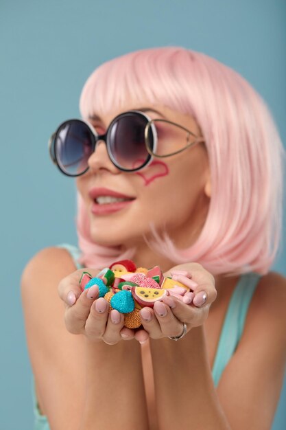 Close up of a flirtatious female model on blue background wearing a pink wig holding different kinds of candies Pretty glamorous woman pink hair charm sweets lifestyle enjoyment sugar