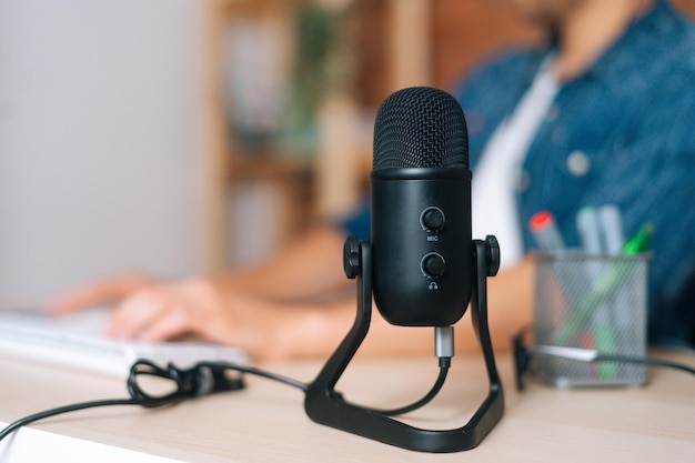 Close-up focus on foreground of black professional studios microphone standing at wooden table, unrecognizable business man working on laptop computer on blurred background.