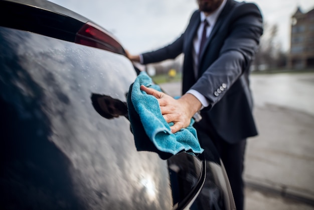 Close up focus hand view of stylish bearded handsome young\
hardworking man in suit cleaning window with a blue microfiber\
cloth on the manual self-service car washing station.