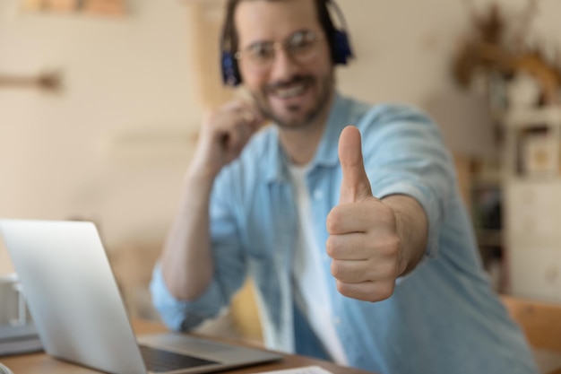 Foto close-up focus van lachende jongeman cliënt in koptelefoon toon duim omhoog geef aanbeveling aan online cursus gelukkige tevreden mannelijke klant aanbevelen goede kwaliteit verre training of opleiding