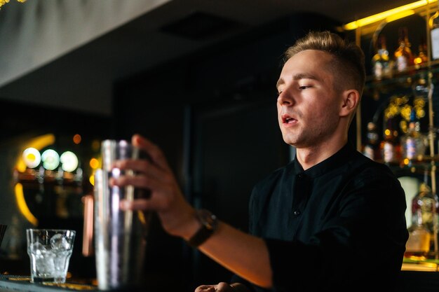 Primo piano di un barista concentrato che prepara cocktail alcolici rinfrescanti in piedi dietro il bancone del bar in una moderna discoteca buia, su sfondo sfocato di scaffali con diverse bevande alcoliche.