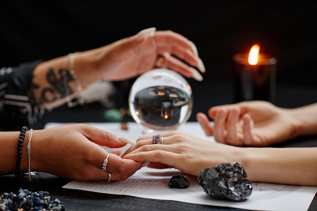 Close up of fortune teller holding hand of young woman during\
spiritual seance with crystal ball