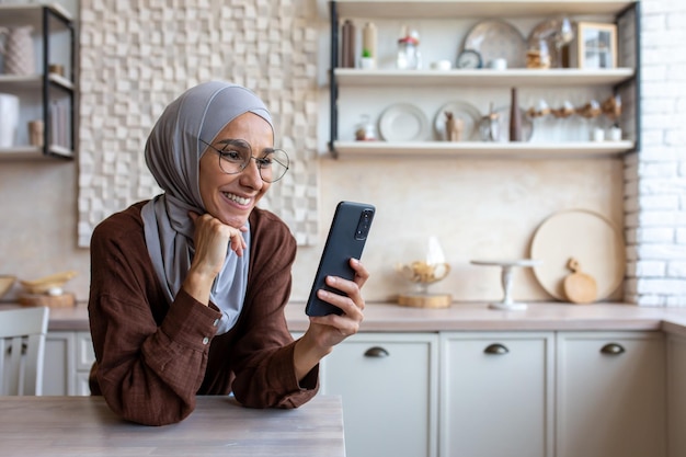 Close-up foto lachende jonge moslimvrouw in hijab die thuis in de keuken staat en mobiel gebruikt