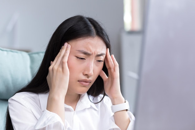 Close-up foto-uitputting op het werk een jonge aziatische vrouw houdt haar hoofd vast en voelt pijn grimassen zitten