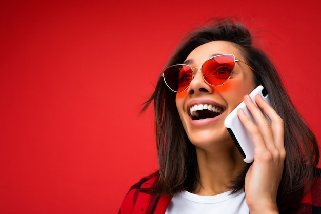 Close-up foto van aantrekkelijke gelukkig positieve jonge brunet vrouw dragen stijlvolle rode shirt wit t-shirt en rode zonnebril geïsoleerd op rode achtergrond communiceren op mobiele telefoon opzoeken