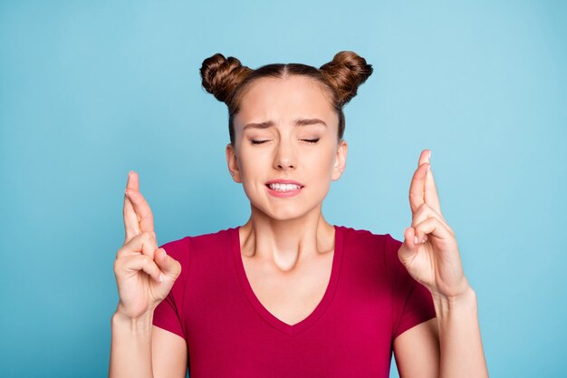 Foto close-up foto van bezorgd onrustig gestrest meisje met gekruiste vingers verwacht vreselijke resultaten van haar examen met gesloten ogen, gekleed in een rood t-shirt geïsoleerd over een blauwe pastelkleurige muur