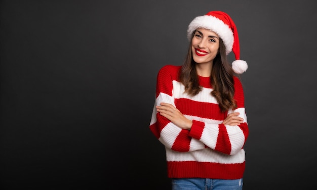 Close-up foto van een aantrekkelijke gelukkige jonge brunette vrouw in kerstmuts met gekruiste handen en poseren geïsoleerd op een grijze achtergrond