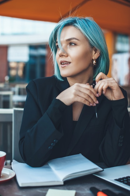 Close-up foto van een blanke vrouw met blauw haar na te denken over iets belangrijks tijdens het gebruik van een laptop en een boek in een cafetaria