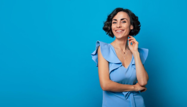 Close-up foto van een charmante gezonde vrouw in een blauwe jurk die in de camera kijkt en glimlacht terwijl ze staat en poseert geïsoleerd op blauwe achtergrond