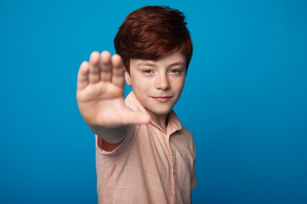 Close-up foto van een lachende jongen met rood haar en sproeten stopbord gebaren op een blauwe muur