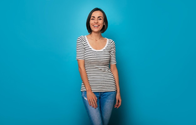 Close-up foto van een mooie glimlachende zelfverzekerde jonge brunette vrouw poseert in een gestreept t-shirt en geïsoleerd op blauwe achtergrond