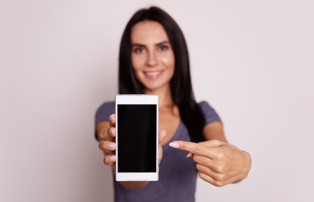 Close-up foto van een schattig meisje met lang zwart haar, die poseert met een smartphone in haar rechterhand en ernaar wijst