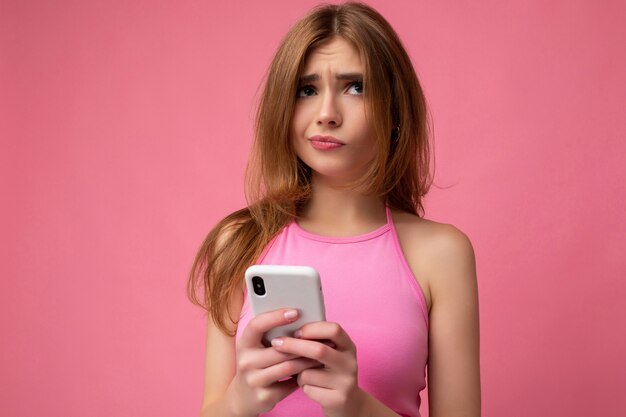 Close-up foto van mooie doordachte jonge blonde vrouw, gekleed in roze top evenwicht geïsoleerd op roze