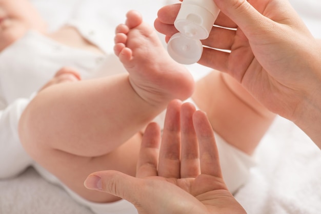 Close-up foto van pasgeboren baby's benen voeten en moeders handen met witte crème tube op geïsoleerde witte achtergrond