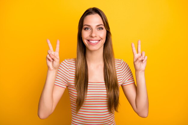 Close-up foto van verbazingwekkende aantrekkelijke dame met v-teken symbolen gedag zeggen tegen vrienden goed humeur dragen casual gestreept t-shirt geïsoleerde gele kleur muur