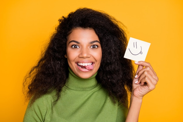 Close-up foto van vrolijke schattige mooie vrij positieve vriendin glimlachend toothily steekt tong uit met papier met haar emoji geïsoleerd levendige kleuren achtergrond