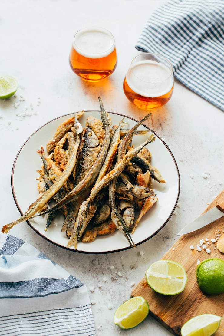 Close-up of fried fish anchovy in a plate next to beer in glasses and lime on a board