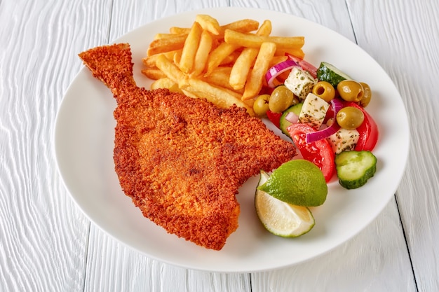 Close-up fried flounder in breadcrumbs served with fresh vegetables, feta, olives greek salad and french fries on a white plate on a wooden table, view from above