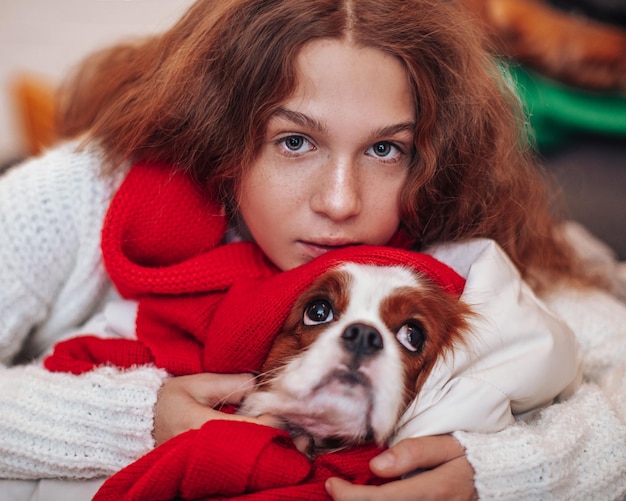 Close up front view of young teen girl with red hair in white sweater and red scarf holding and hugging dog in hands lying on the floor