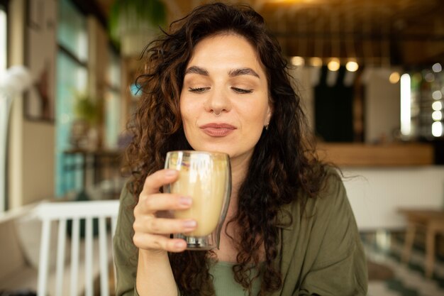 巻き毛の若いきれいな女性の面白い幸せそうな顔を閉じてコーヒーショップでコーヒーを飲んで楽しんでいます