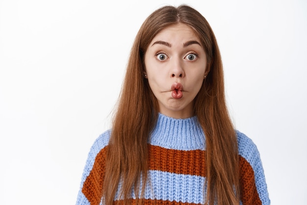 Close up of funny surprised girl, pucker lips and stare with interest and excitement at front, standing in sweater against white wall