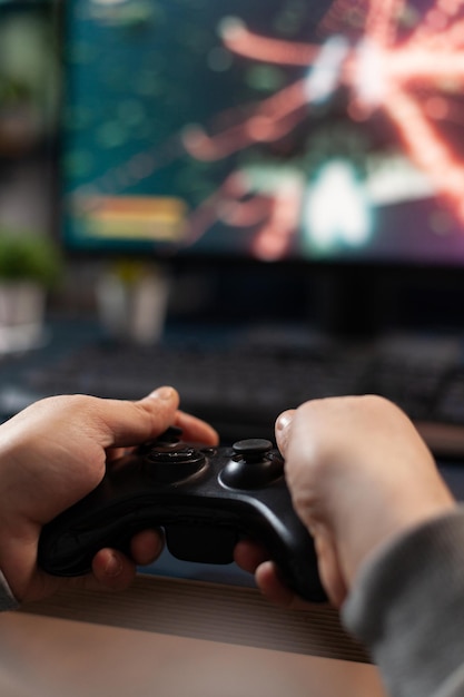 Close up of gamer holding controller to play video games in front of computer. Player using joystick and playing online games on monitor, sitting at desk. Man gaming with modern equipment.