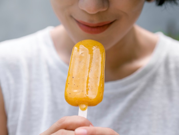 Close-up gele ijslolly in de hand van een mooie, gelukkige Aziatische vrouw met een casual wit mouwloos shirt, buitenshuis. Vrouw die ijslollys eet. Glimlachend vrouwtje genieten van ijslolly in de zomer.