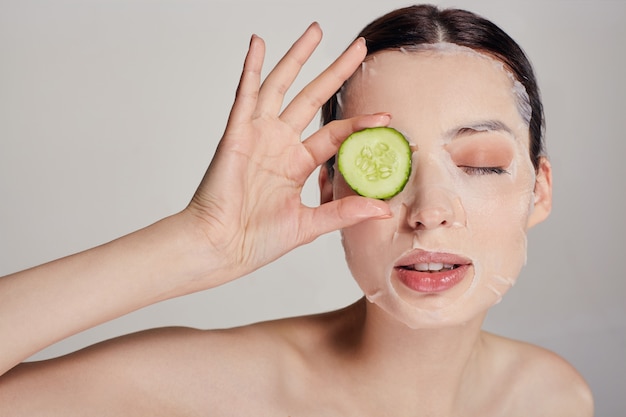 Close up gentle sophisticated calm woman in a moisturizing mask with a fresh cucumber on the face in the serious with close eye and right hand holds a cucumber near eye