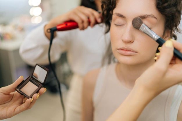 Close-up gesneden shot van onherkenbare vrouwelijke make-up artist toepassen highlighter voor gezicht sculpting van mooie jonge vrouw in moderne schoonheid studio Hairstylist doen stijlvolle kapsel aan langharige dame