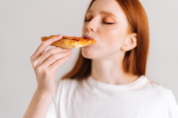 Close-up gezicht van gelukkige aantrekkelijke jonge vrouw met gesloten ogen eetlust heerlijke pizza eten staande op een witte geïsoleerde achtergrond. mooie roodharige vrouw die smakelijke maaltijd eet, selectieve aandacht.