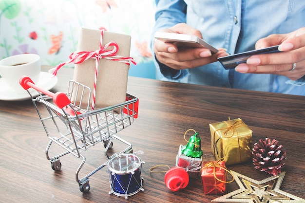 Close up of gift box and Christmas decorations in shopping cart with woman hand online paying in background