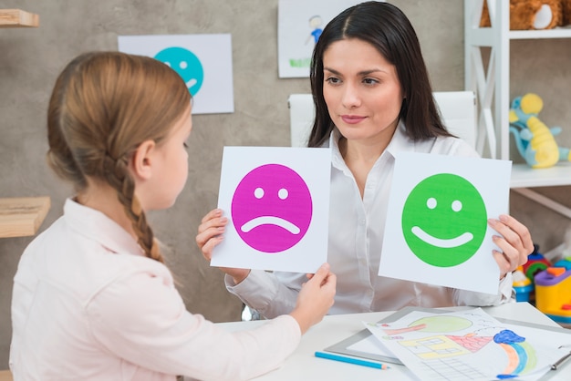 Photo close-up of a girl choosing sad face emoticons paper held by smiling young psychologist