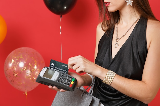 Close up of girl hold wireless modern bank payment terminal to process acquire credit card payments, packages bags on red background air balloons. Happy New Year birthday mockup holiday party concept.
