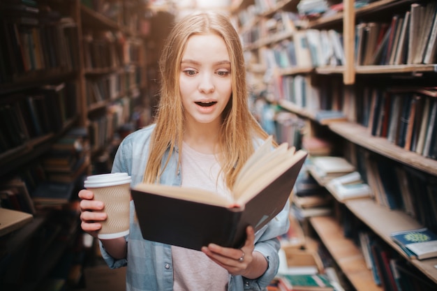 Close up of a girl reading a book and drinking coffee. The book is so interesting she can't stop reading it. The girl is standing in the labrary.