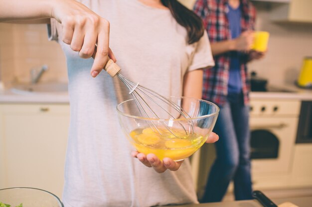 Close up of girl's hand blending eggs with special equipment in glass bowl. Guy stands behind her and holds yellow cup.