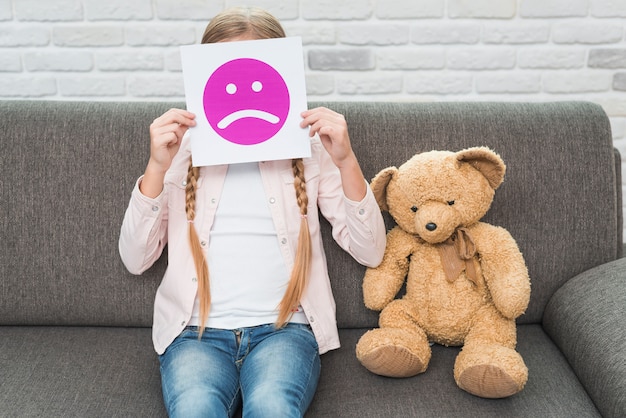 Foto primo piano della ragazza che si siede con teddybear che tiene la carta triste emoticon faccia davanti al viso