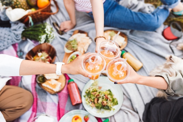 Close up girls holding cup to go with orange juice on beautiful picnic with variety of tasty food in park