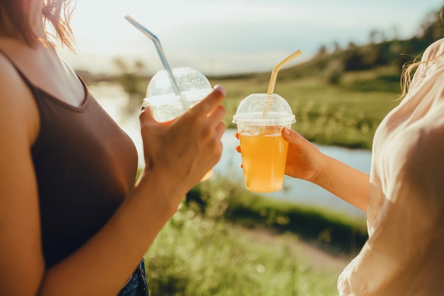 Close up. girls with two plastic cups with orange juice, with straw, at sunset, positive facial expression, outdoor