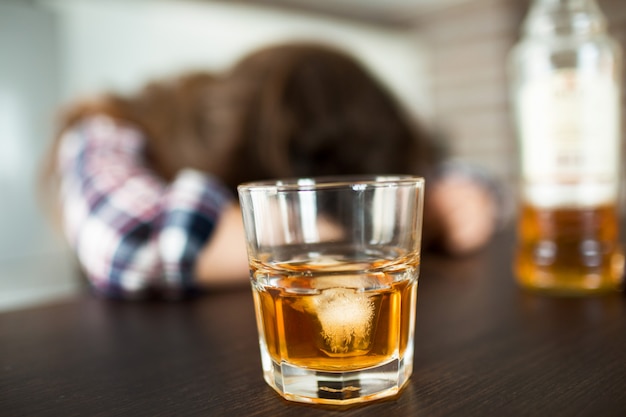 Close up of glass and bottle with whiskey inside. Drunk woman sleeping.