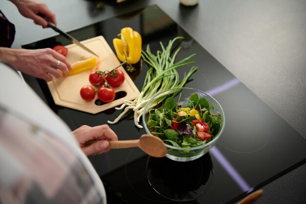 Close-up of a glass bowl with fresh raw salad and male hands\
slicing yellow bell pepper and tomatoes on a cutting board and\
pregnant woman\'s hands holding wooden spoon for mixing raw vegan\
ingredients