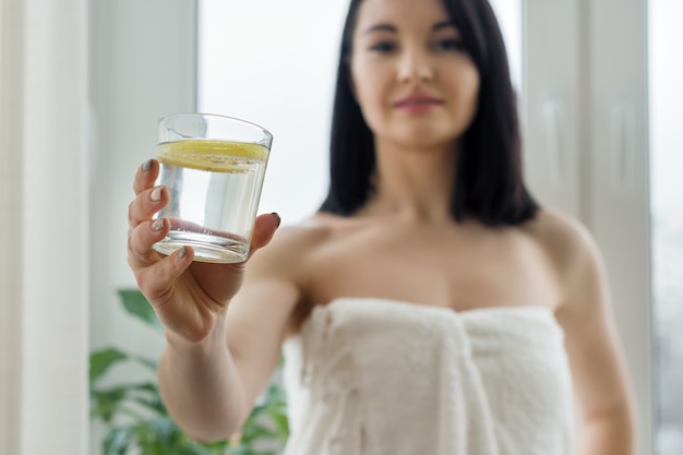 Primo piano di acqua di vetro con il limone a disposizione della giovane donna che si leva in piedi a casa vicino alla finestra.