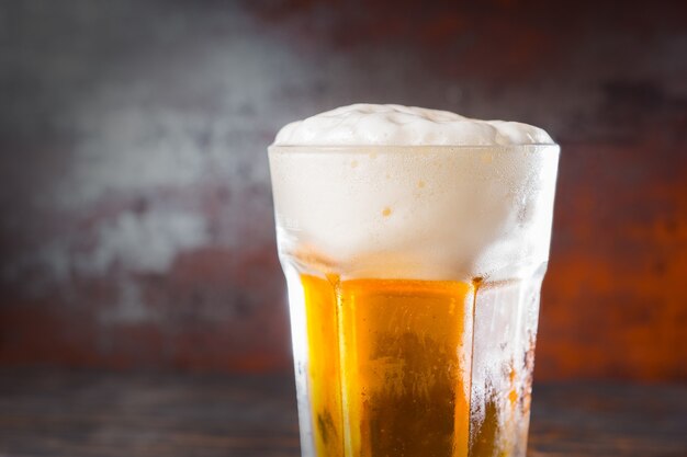 Close up of glass with a light beer and a large head of foam on old dark desk. Drink and beverages concept