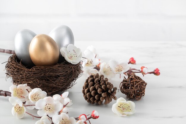 Close up of golden and silver Easter eggs in the nest with white plum flower on bright white wooden table background.