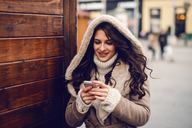 Close up of gorgeous Caucasian woman dressed in coat using smart phone for reading or writing message while standing outdoors on cold weather.