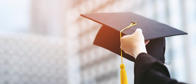 close up graduate hand hold show hat in background school building. shot of graduation cap during commencement university degree , education student success learning concept.