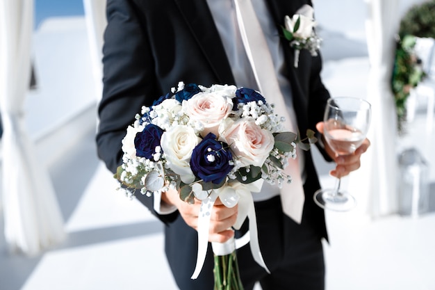 Close-up of the groom in an elegant suit with a bouquet of the bride and a glass of light wine, selective focus