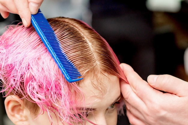 Close-up hairdresser hands are separating the pink hair of young woman with comb in hair salon.